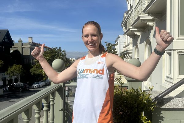 Woman stands on a balcony giving a thumbs up sign wearing a LuMind IDSC singlet.